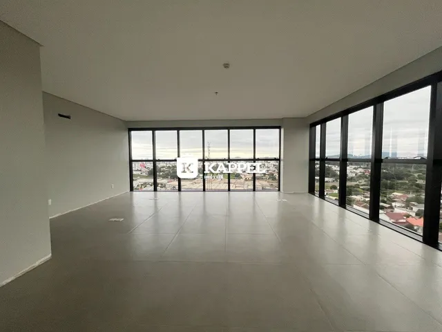 Uma sala ampla com piso claro e grandes janelas que oferecem uma vista da paisagem urbana.