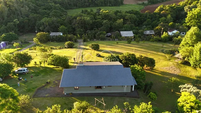 Vista aérea de uma casa simples em uma área verde com campo ao fundo.