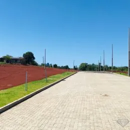 Vista externa de uma área pavimentada com terra vermelha e grama sob um céu azul.