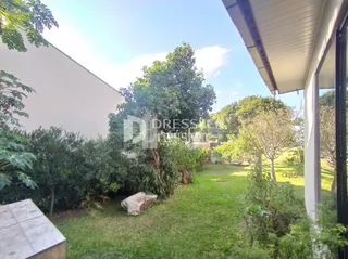 Vista externa de um terreno gramado com vegetação ao redor de uma edificação branca.