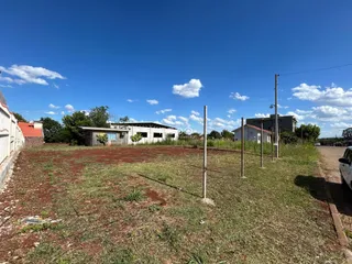 Vista externa de um terreno com vegetação rala e estruturas ao fundo.