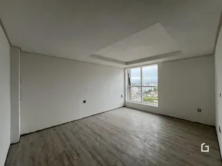 Ambiente interno de uma sala ampla com piso de madeira clara e grande janela.