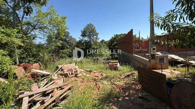 Vista externa de um terreno em construção com vegetação e entulho no primeiro plano.