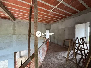 Vista interna de um ambiente em construção com vigas de madeira expostas e piso de cascalho.