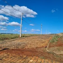 Vista externa de terreno com solo avermelhado e postes de eletricidade sob um céu azul com nuvens.
