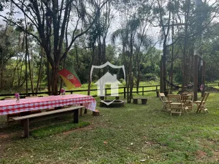 Área externa com mesa e bancos cobertos em um jardim arborizado.