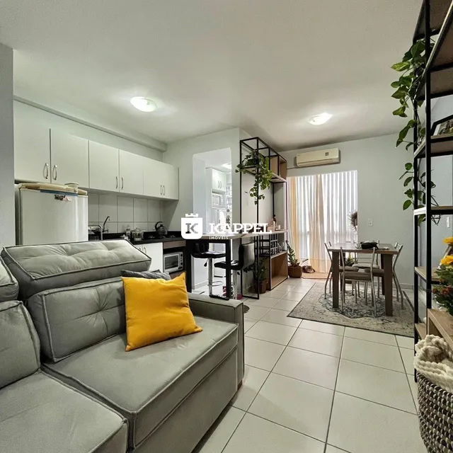 Sala de estar com sofá cinza e detalhes em amarelo, com cozinha moderna ao fundo.