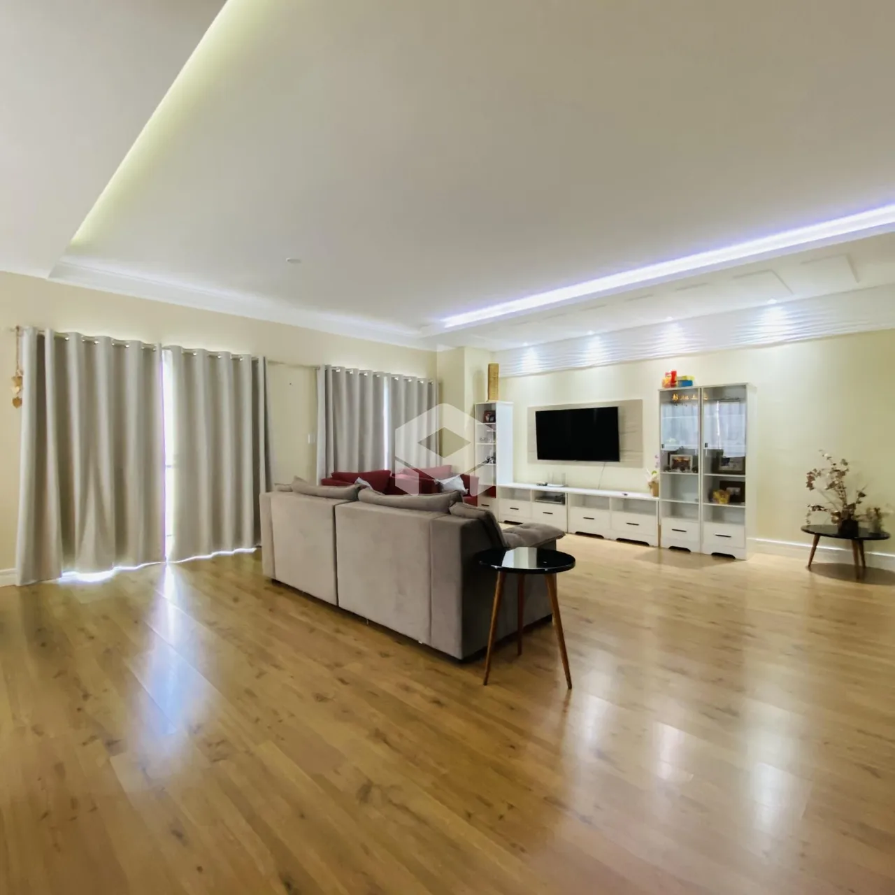 Sala de estar com piso de madeira clara e grandes janelas cobertas por cortinas.