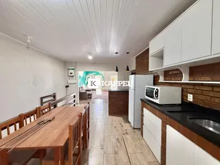 Uma sala de jantar com mesa de madeira e área de cozinha moderna visível no fundo.