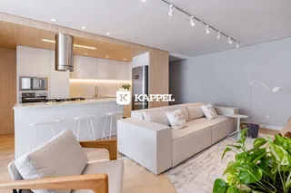 A sala de estar com cozinha aberta apresenta um sofá claro e armários brancos.