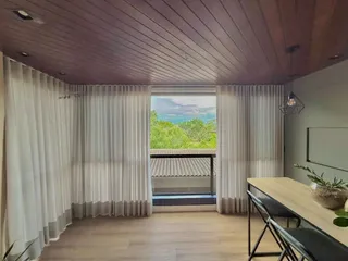 Sala de estar com piso de madeira e cortinas brancas que dão vista para o exterior.