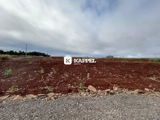 Foto de um terreno aberto com solo avermelhado e vegetação rasteira.