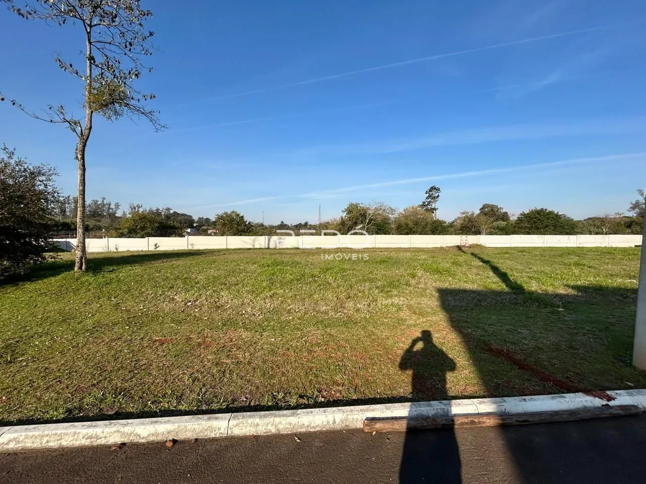 Campo gramado cercado por uma cerca branca e árvores sob um céu azul.